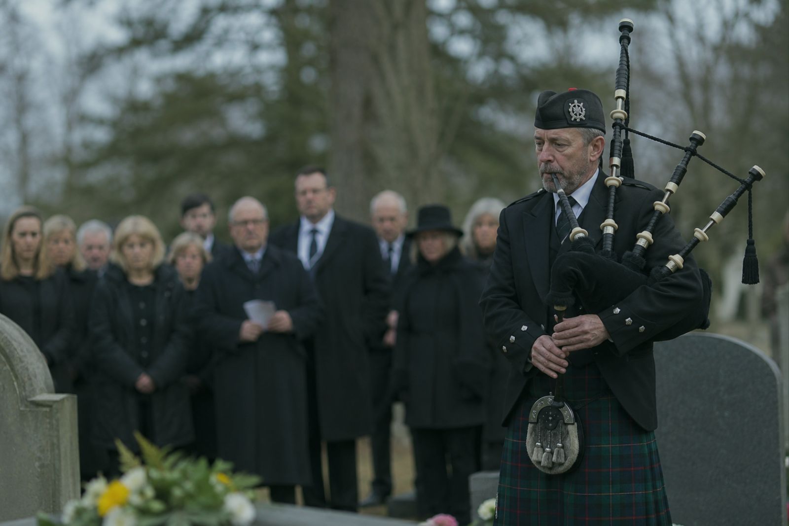 A piper in tartan kilt and black jacket playing the Great Highland Bagpipe in a cemetery, with a group of mourners in the background and gravestones in the foreground