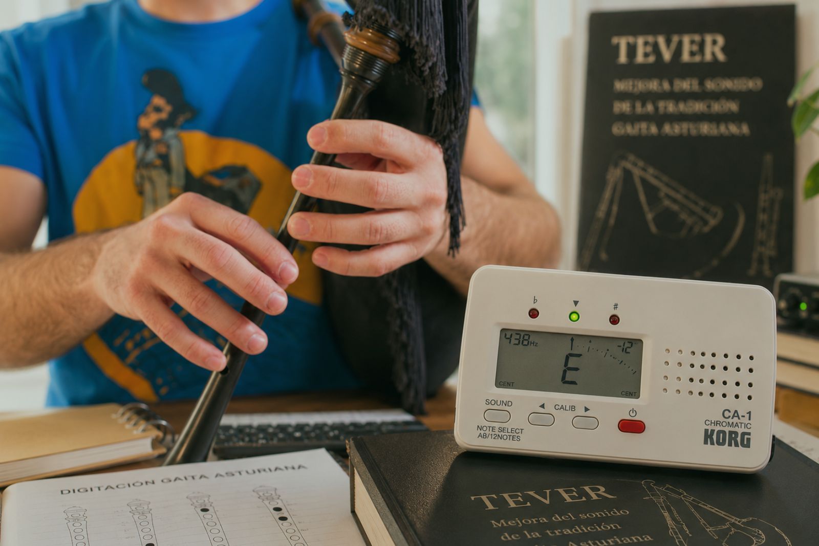Hands holding the chanter of an Asturian bagpipe next to a Korg CA-1 chromatic tuner showing note E, with an open bagpipe fingering notebook and the 'TEVER — Improving the Sound of Tradition — Asturian Bagpipe' poster in the background