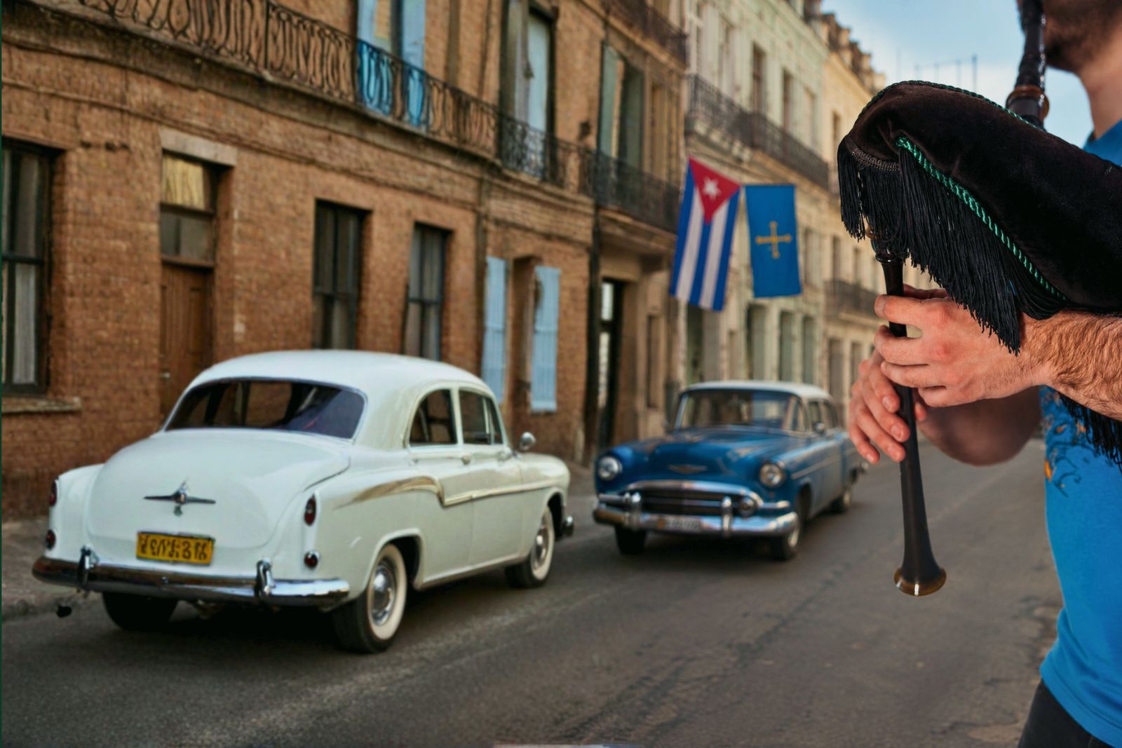 A gaita player playing the Asturian bagpipe on a street in Havana, with classic Cuban cars and the flags of Cuba and Asturias waving together in the background