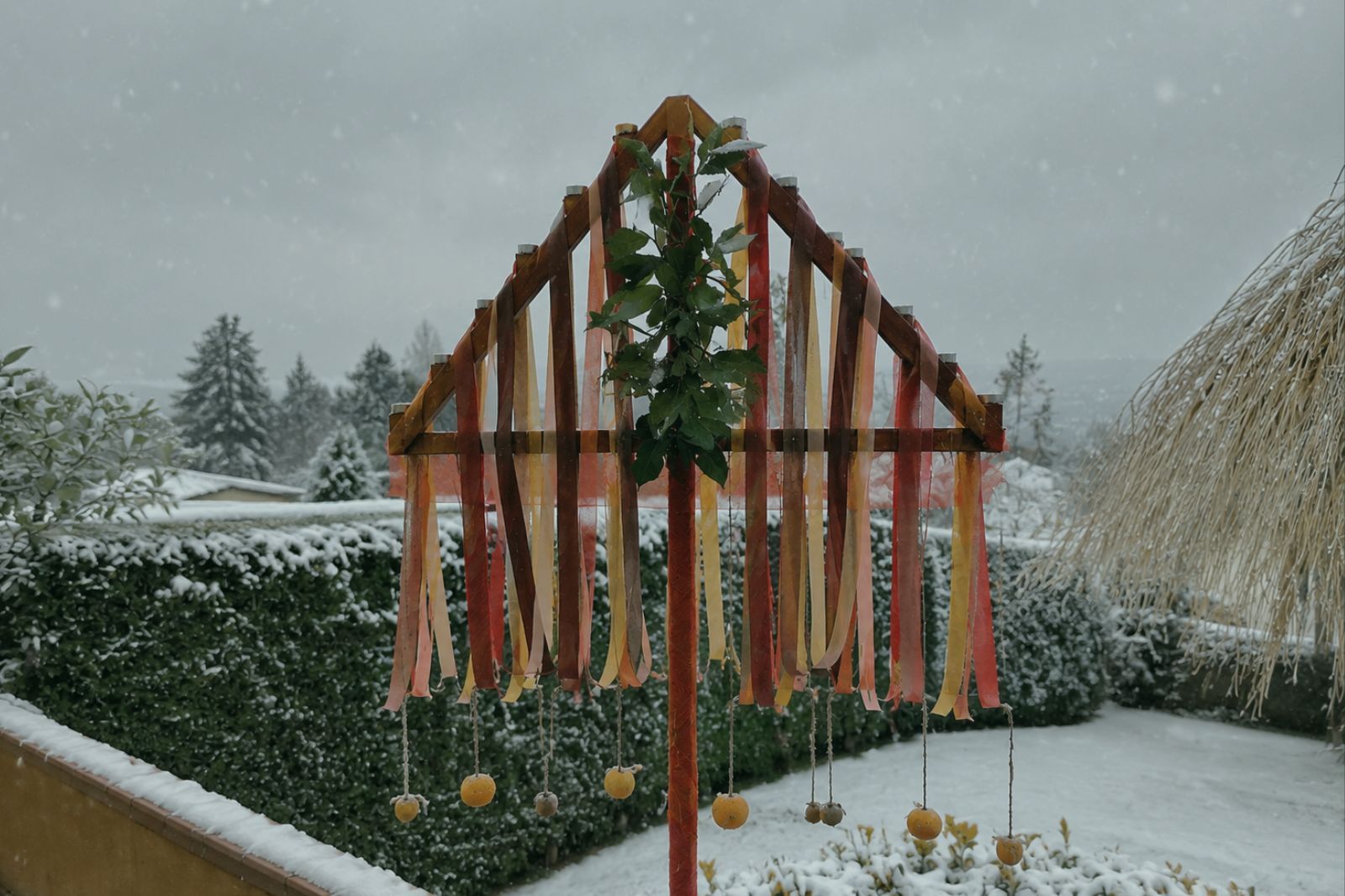 An Asturian Ramu Nadal outdoors over a snowy garden: a triangular wooden frame with coloured ribbons, green branches on the central axis and fruit hanging from the sides