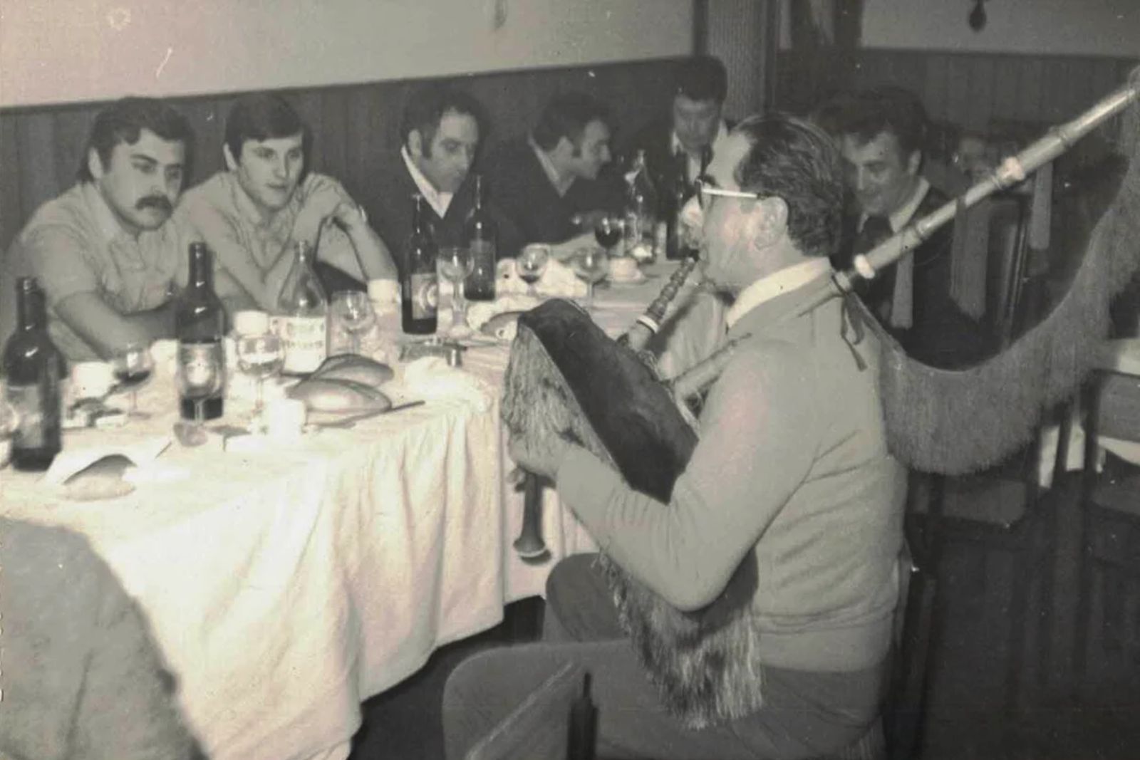 Old black-and-white photograph of a bespectacled piper playing the gaita at a meal, seated beside a table with bottles and glasses, with several men watching him